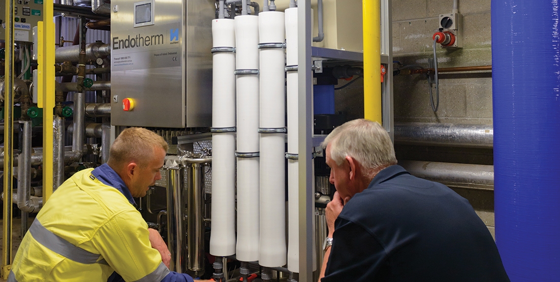 Two service technicians crouched by RO system in hospital plant room, looking at piping of water filtration system