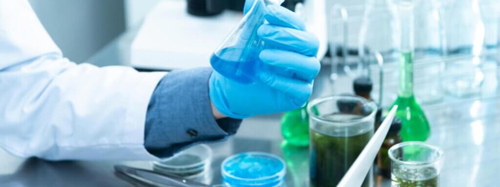 Steps-towards-sustainable-lab_SF Blue and green coloured liquids in beakers and test tubes, with one being held by a gloved hand.